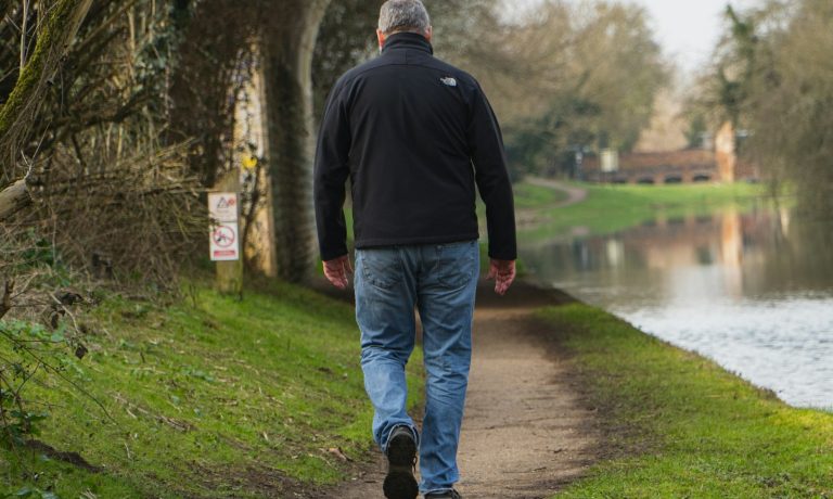 man in black jacket and blue denim jeans walking on pathway near river during daytime
